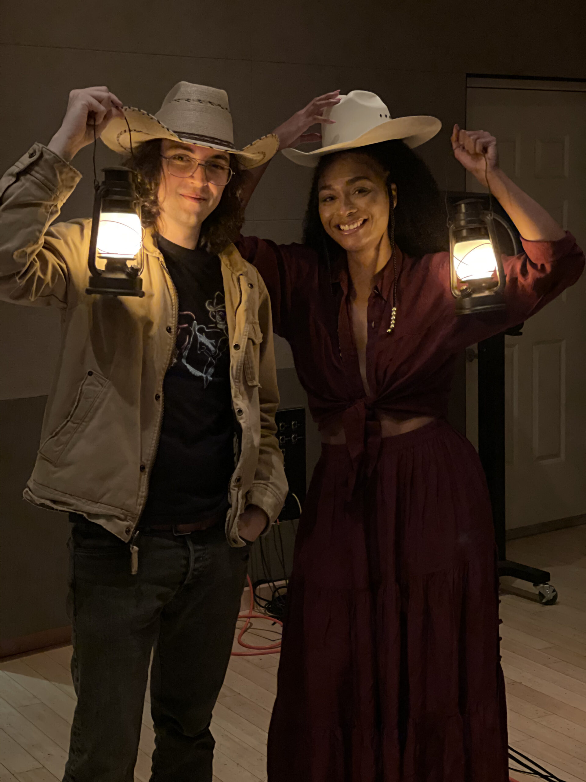 A man and a woman wearing cowboy hats stand in the dark holding lanterns