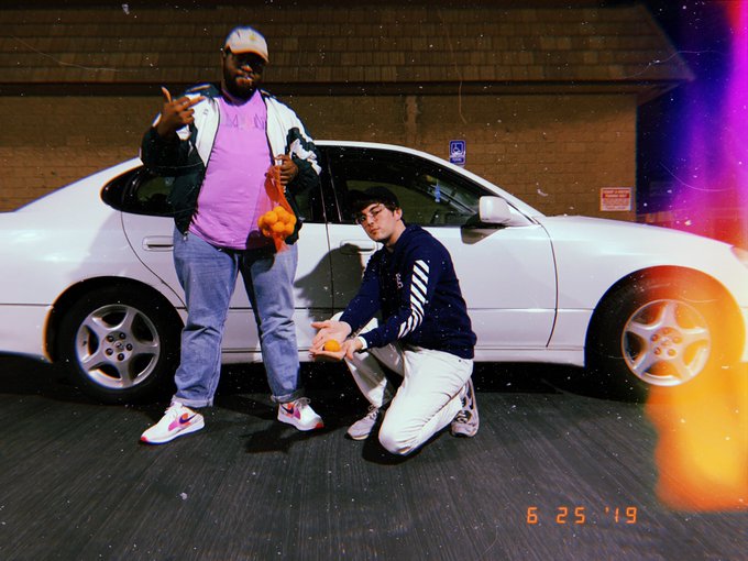 Comedian Jamel Johnson and host Sama'an Ashrawi holding a bag of clementines and posing next to a white mercedes