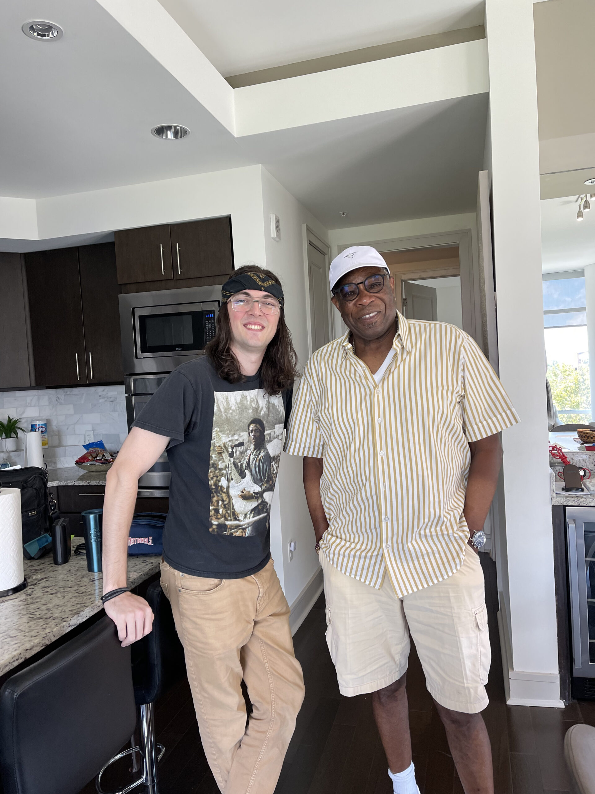 Two men, one younger and fair skinned with long hair and a bandana, the other older and Black with a big grin, pose for a photo in a kitchen.